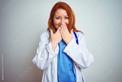 Young redhead doctor woman using stethoscope over white isolated background laughing and embarrassed giggle covering mouth with hands, gossip and scandal concept photo