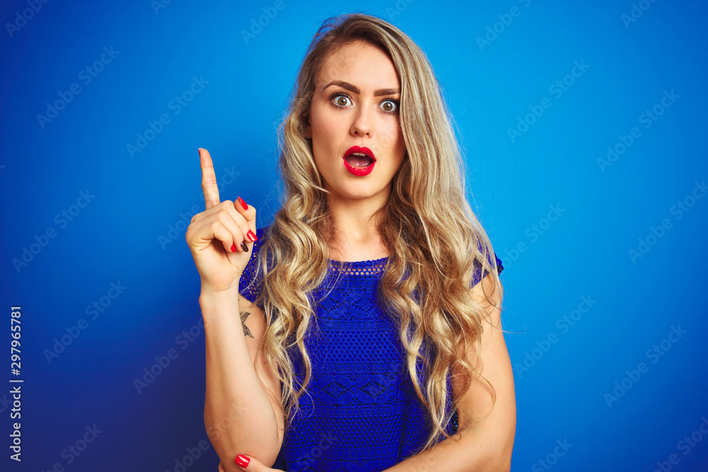 Young beautiful woman standing over blue isolated background pointing finger up with successful idea. Exited and happy. Number one. Young beautiful woman standing over blue isolated background pointing finger up with successful idea. Exited and happy. Number one.