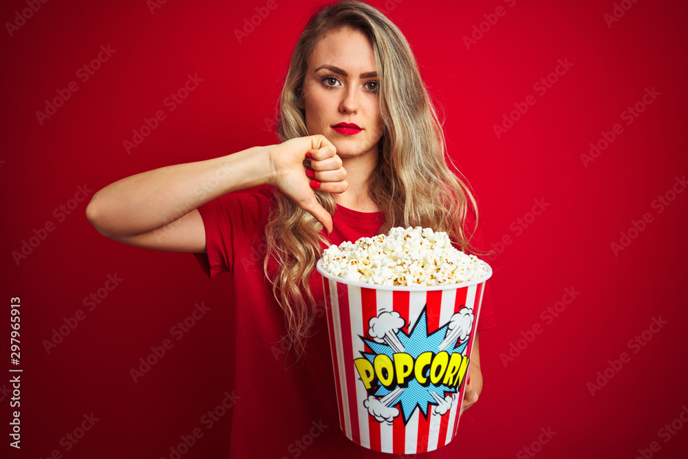 Young beautiful woman wearing t-shirt eating popcorn over red isolated background with angry face, negative sign showing dislike with thumbs down, rejection concept