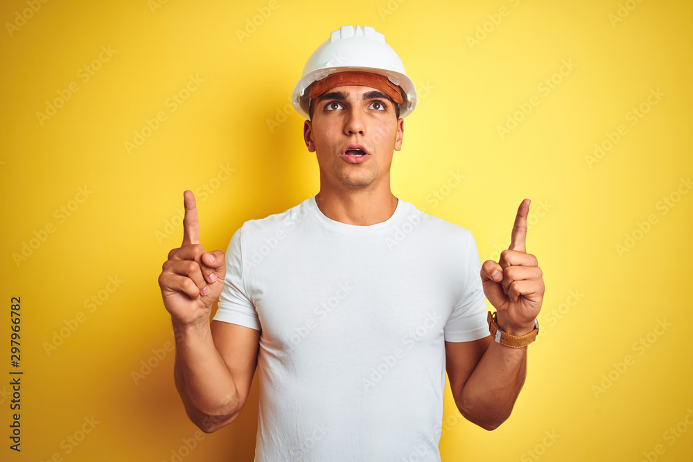 Young handsome man wearing construction helmet over yellow isolated background amazed and surprised looking up and pointing with fingers and raised arms. Young handsome man wearing construction helmet over yellow isolated background amazed and surprised looking up and pointing with fingers and raised arms.