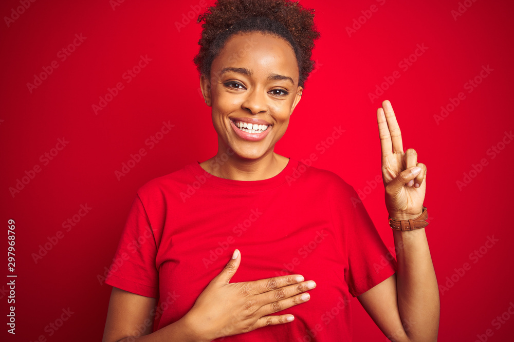 Young beautiful african american woman with afro hair over isolated red background smiling swearing with hand on chest and fingers up, making a loyalty promise oath Young beautiful african american woman with afro hair over isolated red background smiling swearing with hand on chest and fingers up, making a loyalty promise oath