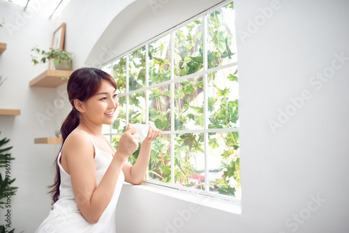 Asian woman in the morning holding a cup of tea or coffee and standing behind the window