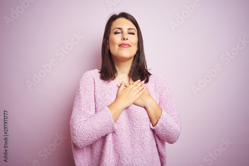 Young beautiful brunette woman wearing a sweater over pink isolated background smiling with hands on chest with closed eyes and grateful gesture on face. Health concept.