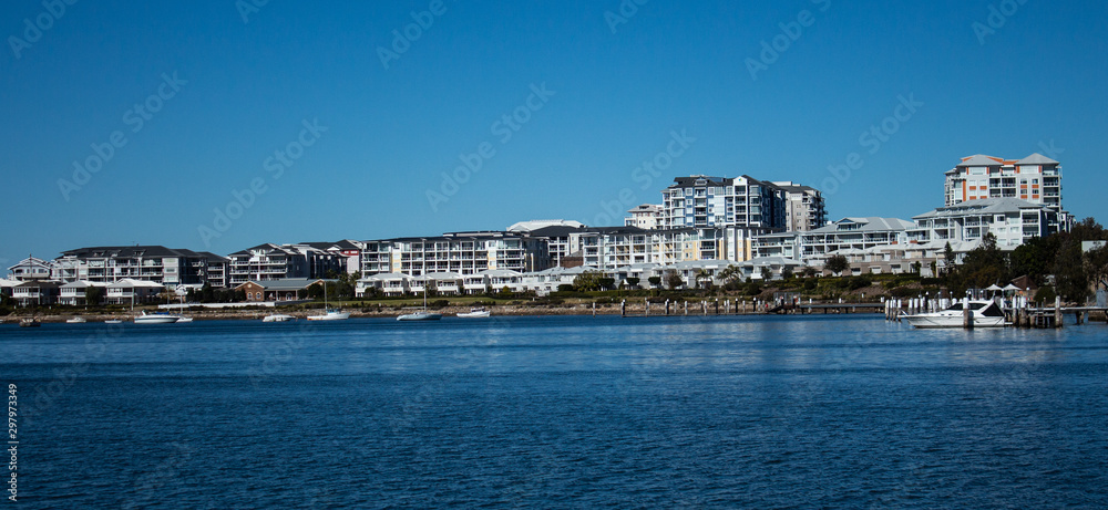 Large waterside houses, apartment condominiums in suburban community on riverfront with boats moored at wharf, blue sky in background