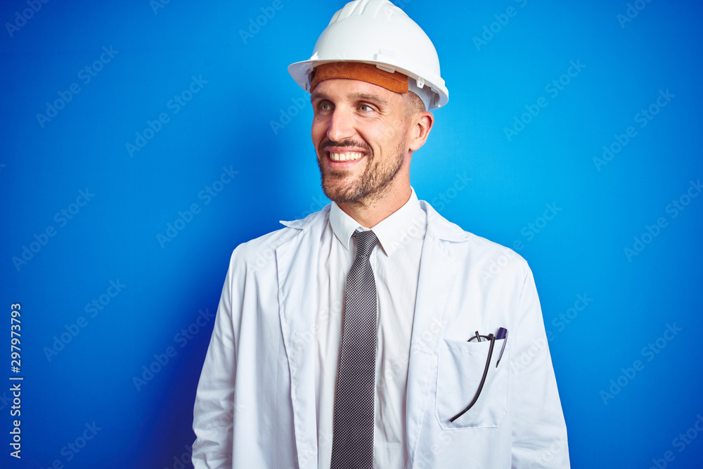 Young handsome engineer man wearing safety helmet over blue isolated background looking away to side with smile on face, natural expression. Laughing confident. Young handsome engineer man wearing safety helmet over blue isolated background looking away to side with smile on face, natural expression. Laughing confident.