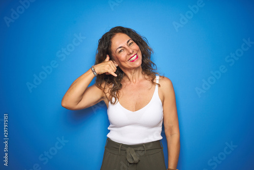 Middle age senior woman with curly hair standing over blue isolated background smiling doing phone gesture with hand and fingers like talking on the telephone. Communicating concepts.