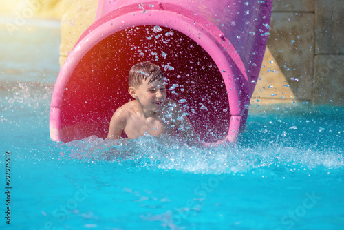 Boy having fun on water slide in the aqua fun park glides, happy falling into water and water splashes are all over. © Artem