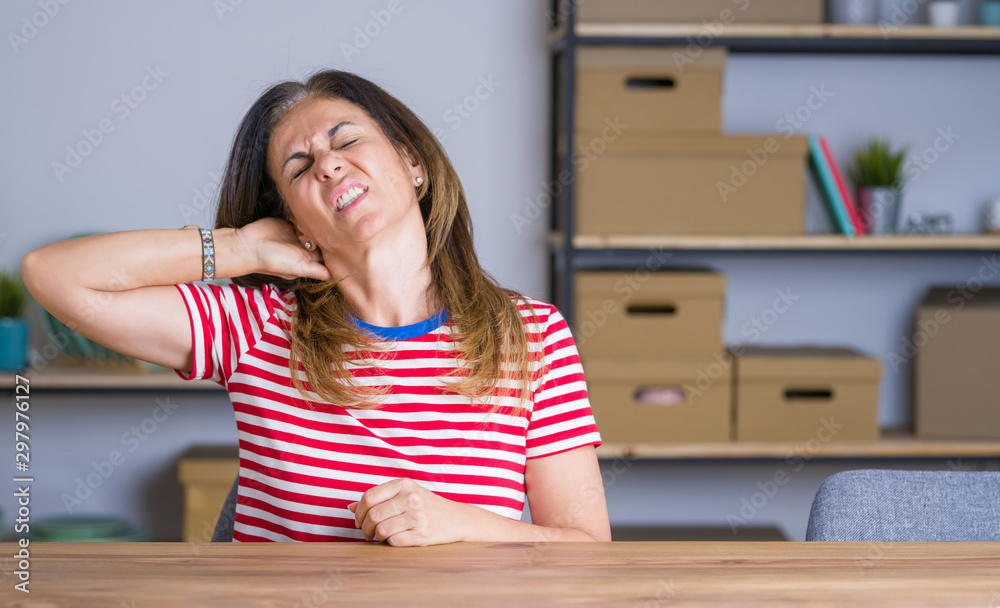 Middle age senior woman sitting at the table at home Suffering of neck ache injury, touching neck with hand, muscular pain Middle age senior woman sitting at the table at home Suffering of neck ache injury, touching neck with hand, muscular pain