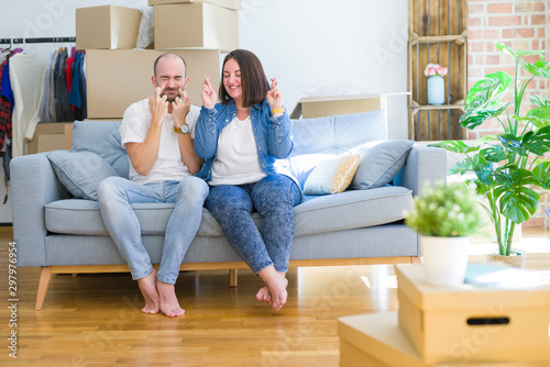 Young couple sitting on the sofa arround cardboard boxes moving to a new house gesturing finger crossed smiling with hope and eyes closed. Luck and superstitious concept.