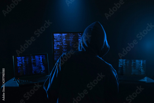 Rear View Of A Hacker Using Multiple Computers For Stealing Data On Desk