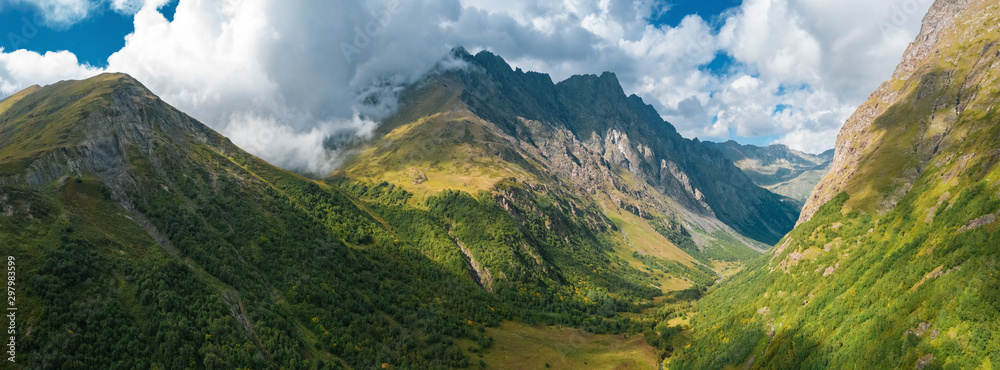 Fototapeta premium Panorama of mountain terrain in early autumn; amazing dramatic clouds on mountain peaks; rivers cutting gorges and canyones in valleys; natural outdoor travel background; sunny day on Caucasus