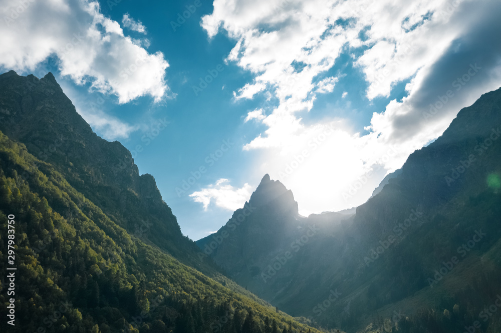 Amazing sunset highland landscape with sun setting on sharp mount peak; natural outdoor travel background; silhouettes of mountain ridges; river valley with last sunrays on overgrown slopes