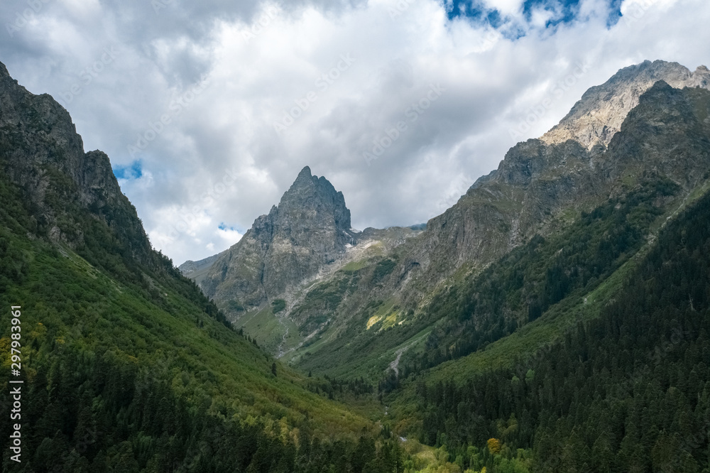Panoramic view of river Malaya Laba valley with mountain ranges on sides, sharp peak Igolchatiy; morning landscape, dramatic cloudy sky, deep pine fir forest; danger narrow gully, steep gradient