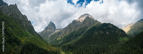 Wide panoramic view of river Malaya Laba valley with mountain ranges on sides, sharp peak Igolchatiy; morning landscape with dramatic cloudy sky, deep pine fir forest; danger stony slopes, Caucasus