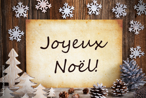 Old Paper With French Text Joyeux Noel Means Merry Christmas. Christmas Decoration Like Tree, Fir Cone And Snowflakes. Brown Wooden Background