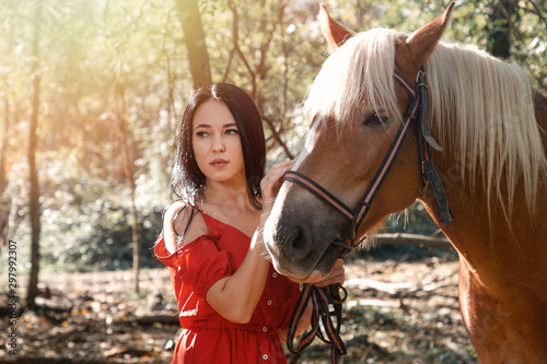 Portrait of Young beautiful brunette woman in red dress posing near horse. Sunlight
