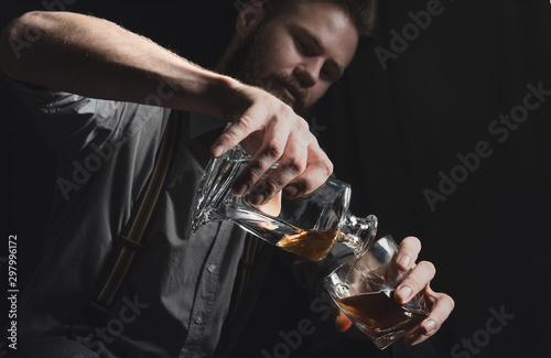 A young man with a beard pours alcohol from a decanter into a glass.