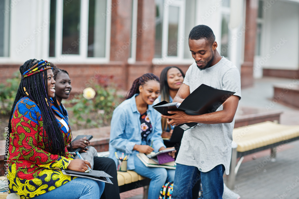 Obraz premium Group of five african college students spending time together on campus at university yard. Black afro friends studying at bench with school items, laptops notebooks.