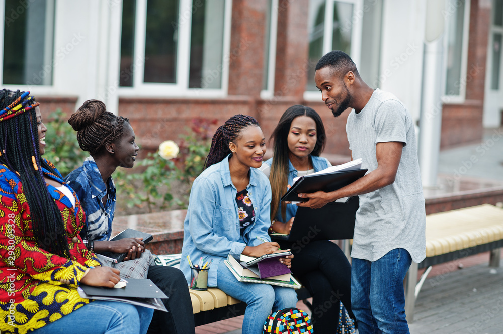 Obraz premium Group of five african college students spending time together on campus at university yard. Black afro friends studying at bench with school items, laptops notebooks.