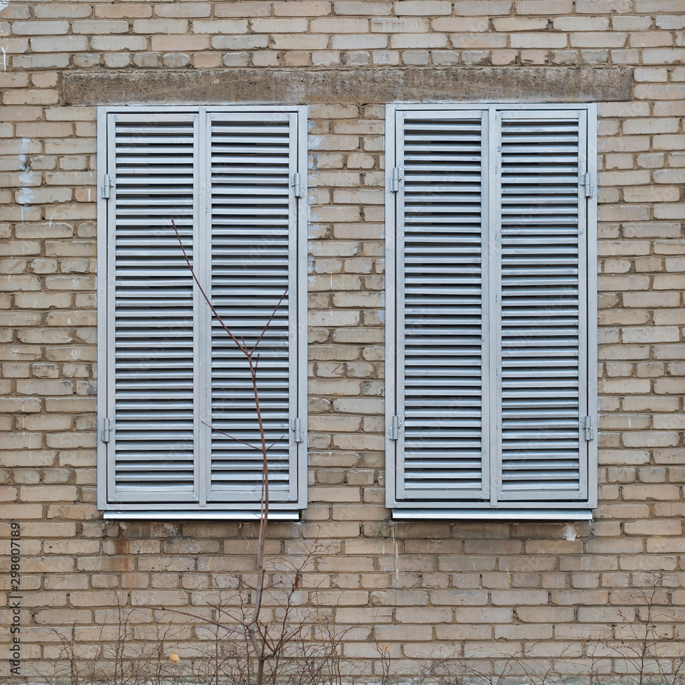 Two windows with closed grey metal shutters on dirty brick wall background
