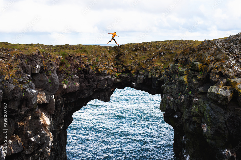 Tourist jumps over a natural rock bridge in Arnarstapi, Iceland