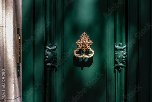Old door with knocker. Green painted wooden framed door with gorgeous antique ornate knocker and carving sculptural details. Architectural features of Paris door in bright sunlight with shadows.