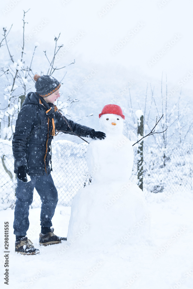  Winter teenager in a jacket and hat sculpts a snowman on a winter background with falling snow in frozen day concept