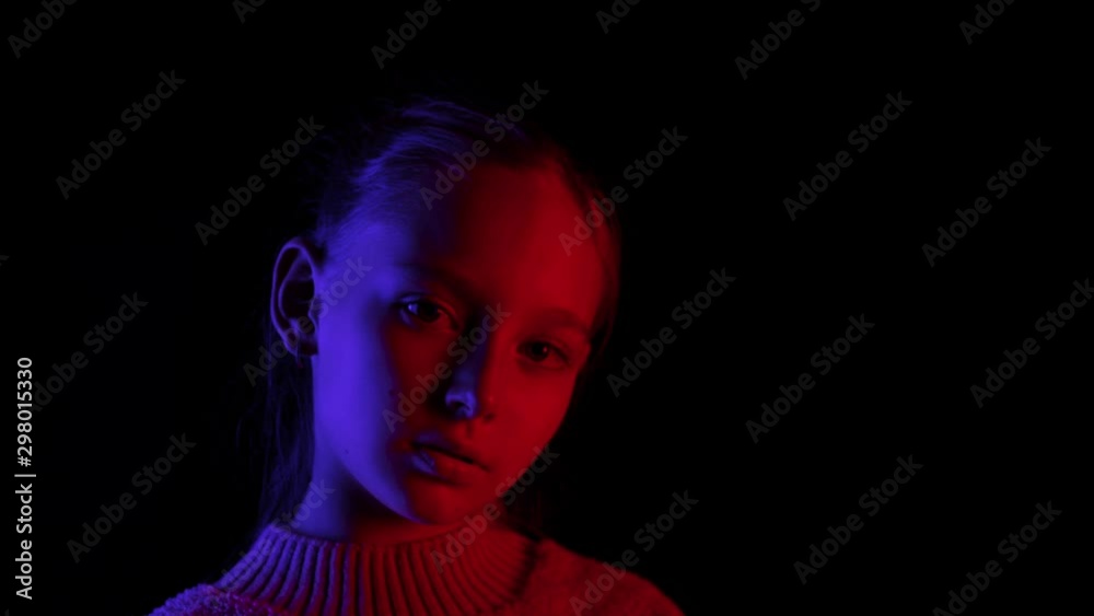Young girl looking up on black background in dark studio with blue and red lighting. Portrait girl teenager posing front camera in black studio with neon lighting background