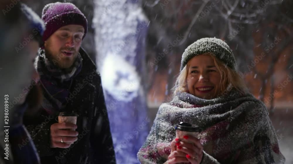 Group of happy friends walking under snowfall with paper coffee cups in hands and have fun playing with snow. People warming up with hot beverages and hanging on winter night. Christmas and New Year.