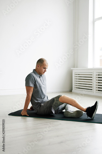 Young man with prosthesis on the leg sitting on the floor on exercise mat and exercising in the room © AnnaStills