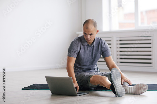 Young man with prosthesis on the leg sitting on the floor on exercise mat and using laptop computer during sports training