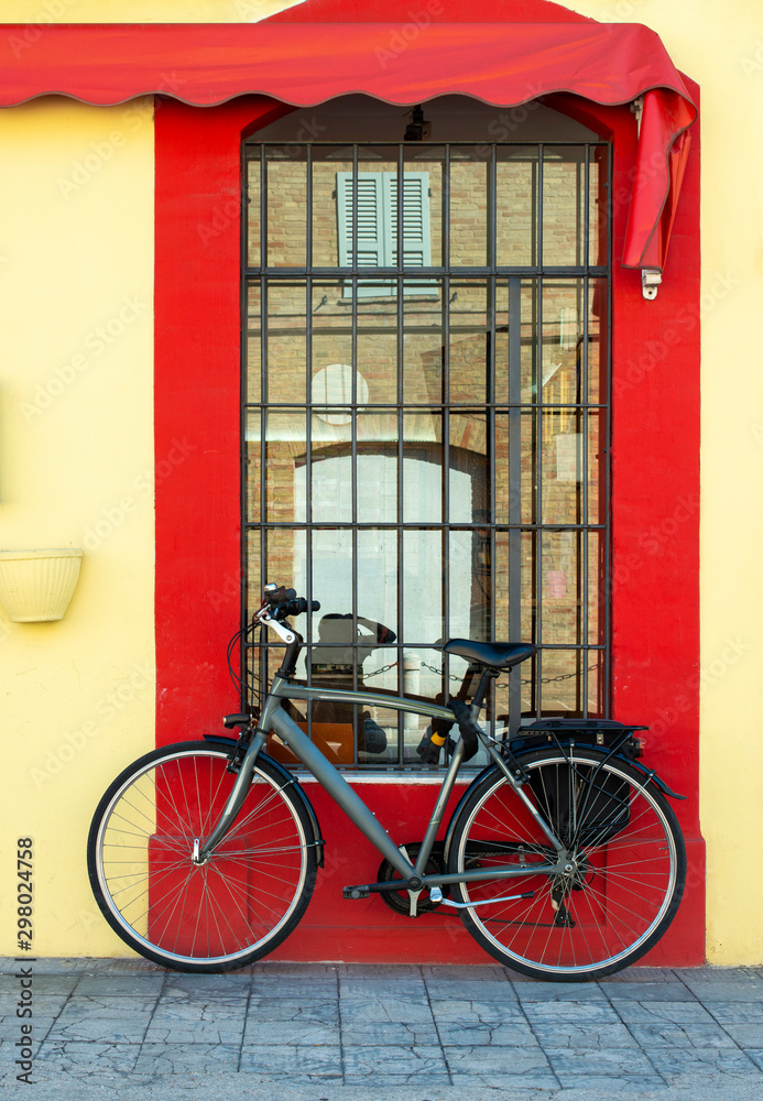Grey bike in front of yellow facade and red window. Bicycle with trunk.