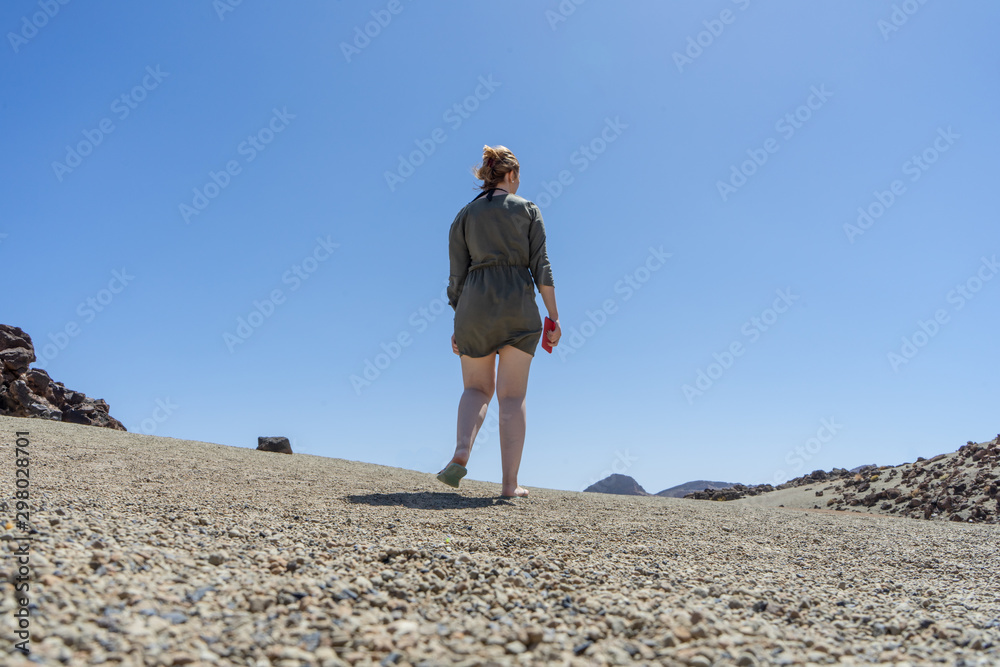 Blond hair teen in relaxing mood at remote lunar, volcanic and arid desert. Back of Spiritual woman in green dress walking alone on volcano path with white legs. Freedom and ethereal exploration.