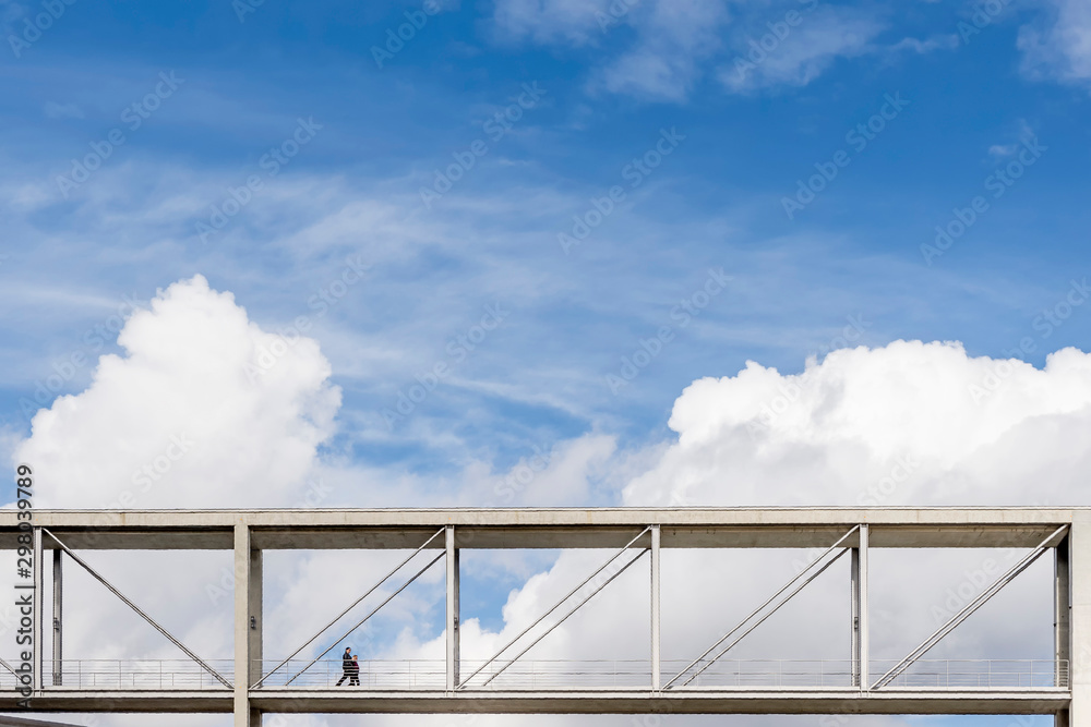 Two people walk on a suspended pedestrian bridge with a dramatic sky in the background in the government district of Berlin, Germany Two people walk on a suspended pedestrian bridge with a dramatic sky in the background in the government district of Berlin, Germany
