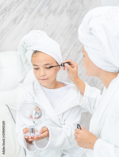 Happy family at home. Mother and young daughter while applying makeup at home. Mom and child girl are in bathrobes and with towels on their heads
