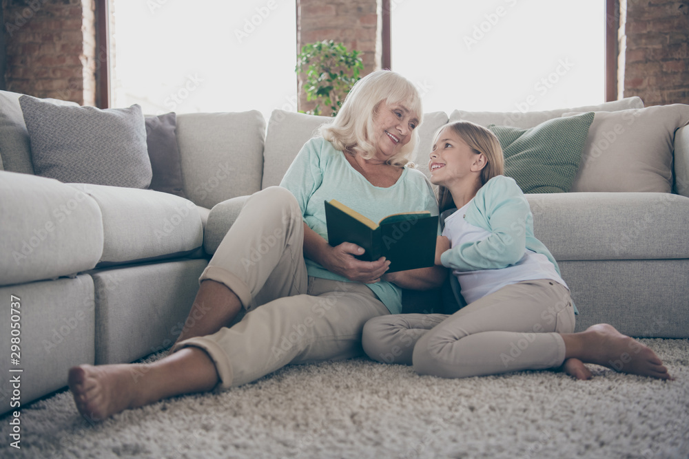 Photo of two people aged granny little granddaughter sitting floor near sofa reading together girl cute hugging grandma listen interested fairy tail house indoors