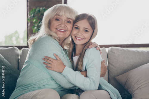Photo of two adorable people cute aged granny little granddaughter sit cozy sofa hug close spending summer weekend holidays together house indoors