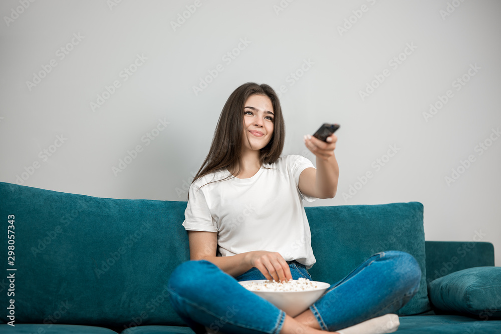 Obraz premium beautiful young woman changes channel while watching TV and eating popcorn feeling comfortable on the sofa at home