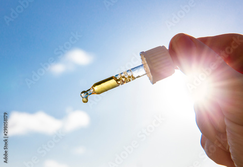 Canvas Print Hand holding dropper pipette with nice golden liquid D-vitamin against sun and blue sky on sunny day