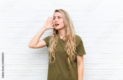 young blonde woman smiling, looking curiously to the side, trying to listen to gossip or overhearing a secret against brick wall background photo