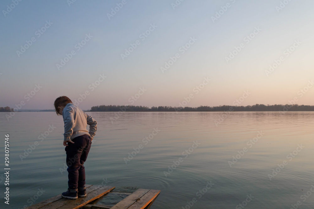 Little kid boy on a wooden pier on a lake, a river looks into the distance at the horizon