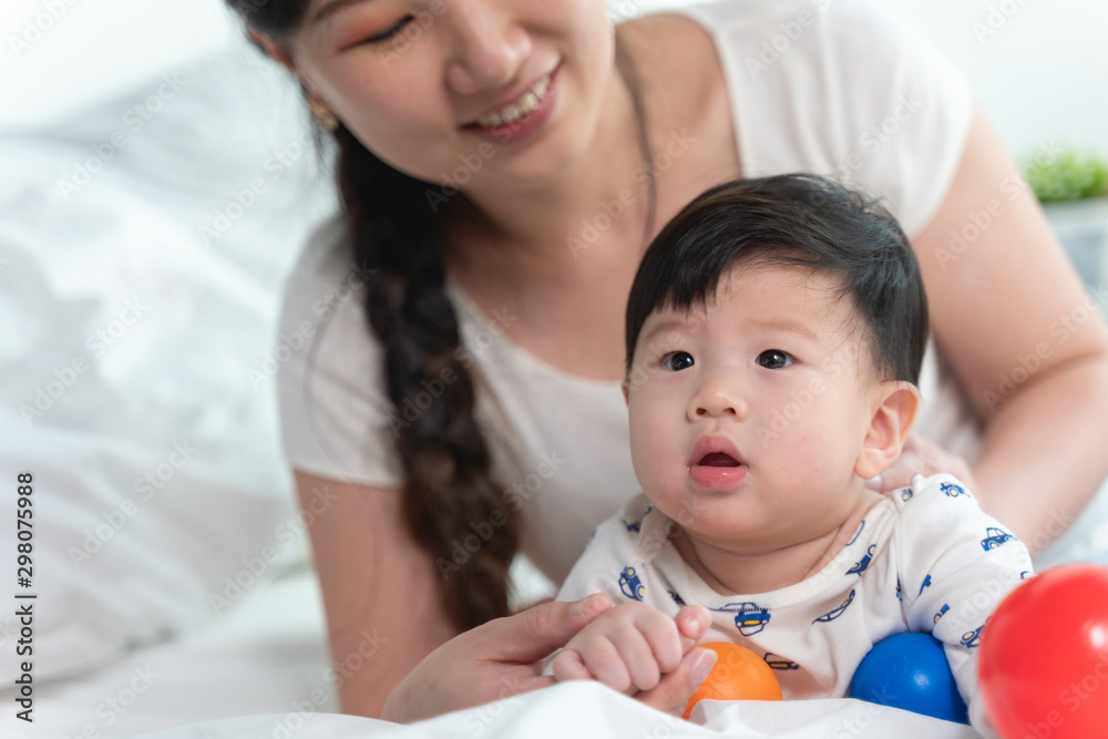 Young beautiful asian mother with asian baby on bed and playing toy ball together on white bed with feeling happy and cheerful and the baby that crawling on the bed.Baby family concept Young beautiful asian mother with asian baby on bed and playing toy ball together on white bed with feeling happy and cheerful and the baby that crawling on the bed.Baby family concept