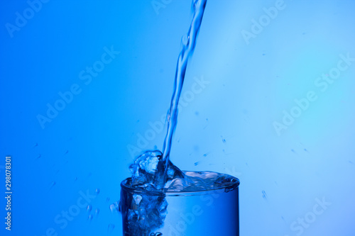 Chorro de agua llenando y rebosando un vaso de agua de cristal o plástico transparente sobre fondo azul