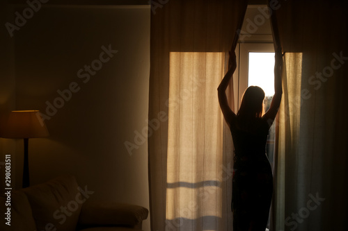 Woman opening curtains in a room, beautiful atmosferic sunny morning