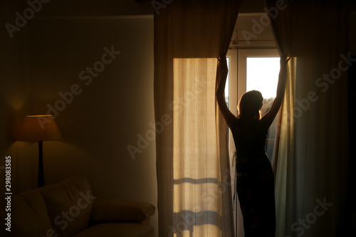 Woman opening curtains in a room, beautiful atmosferic sunny morning