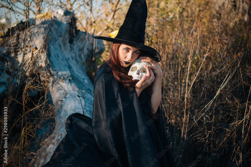 A young witch with pale skin and black lips in a black hat, a dress and a raincoat.  Holds the skull of a dead man. Autumn, fallen tree and tall, dry grass. Halloween, magic, fantasy.