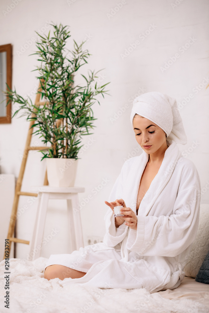 Young beautiful woman in towel and bathrobe applying cream on her hand and face after shower at home