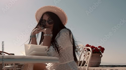  Pretty young brunette with European appearance in a white beautiful dress in black stylish glasses and a hat sits at a table with an open notebook and enjoys the warm weather photo