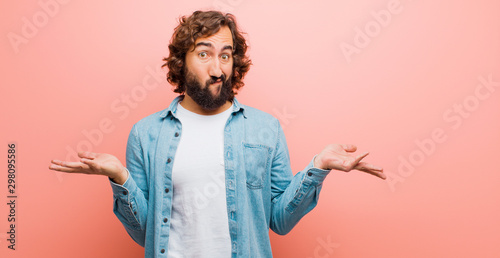 young bearded crazy man feeling puzzled and confused, doubting, weighting or choosing different options with funny expression against flat color wall