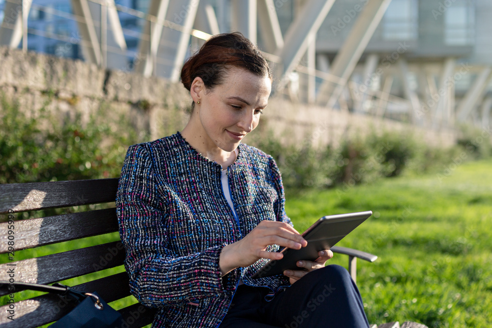 Serious female entrepreneur in elegant jacket frowning and using tablet while sitting on bench on sunny day in city park Serious female entrepreneur in elegant jacket frowning and using tablet while sitting on bench on sunny day in city park